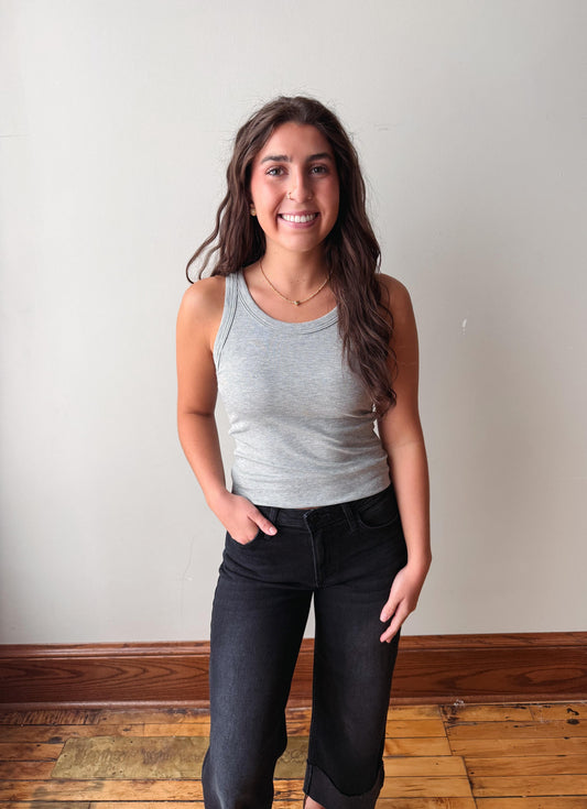 A woman with long brown hair stands indoors wearing a light gray ribbed tank top and dark charcoal jeans, with her hands in her pockets, against a plain off-white wall and wooden floor.