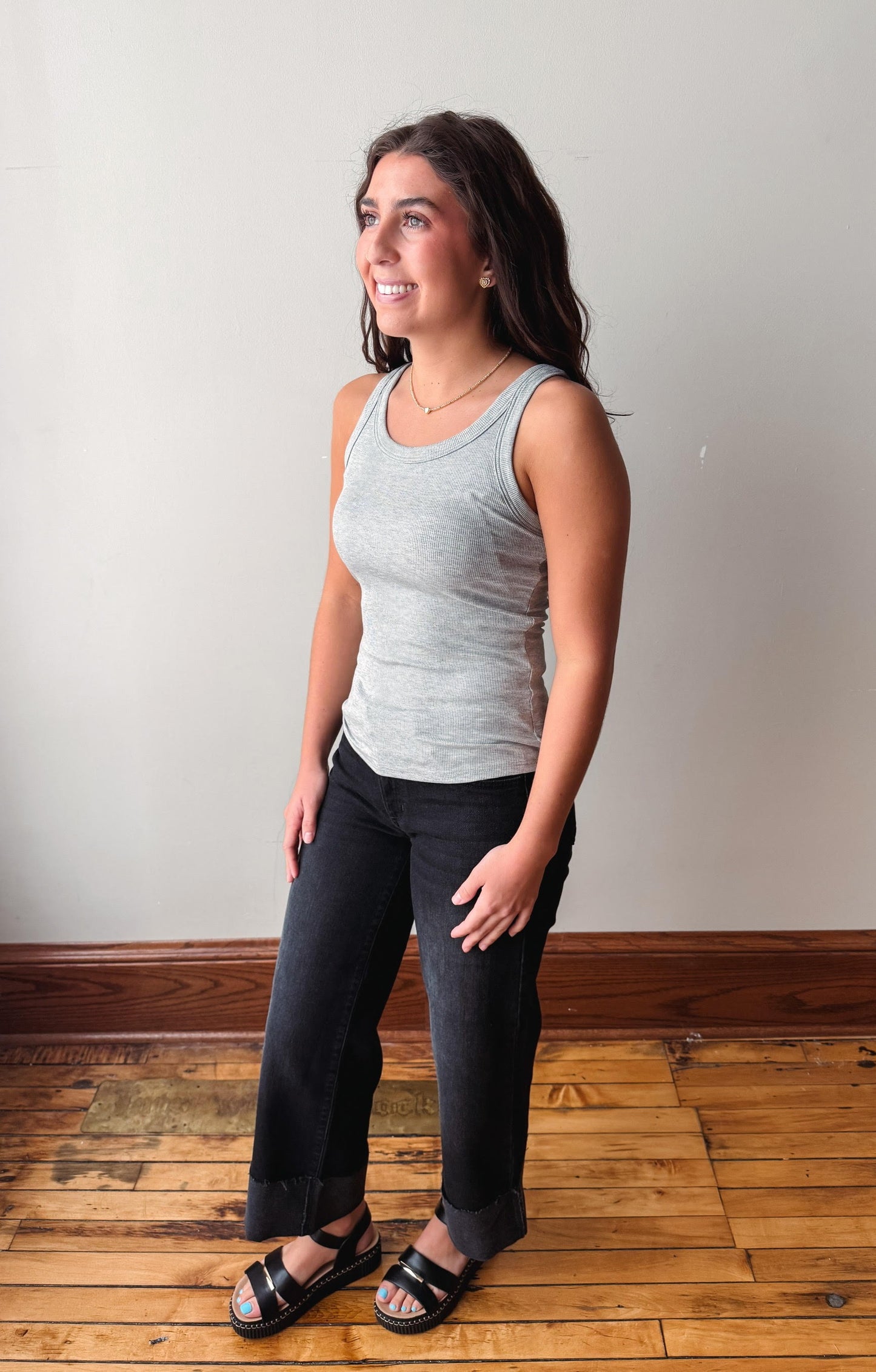 A woman modeling a light gray ribbed tank top and dark charcoal gray wide-leg jeans with black sandals, standing on a wooden floor against a plain off-white wall.