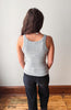 A woman viewed from behind wearing a light gray ribbed tank top and dark jeans, standing on a wooden floor against a plain off-white wall.
