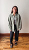 A woman stands in a room with a light-colored wall and wooden floor, wearing a sage green denim jacket over a white top, black pants, and black sandals.