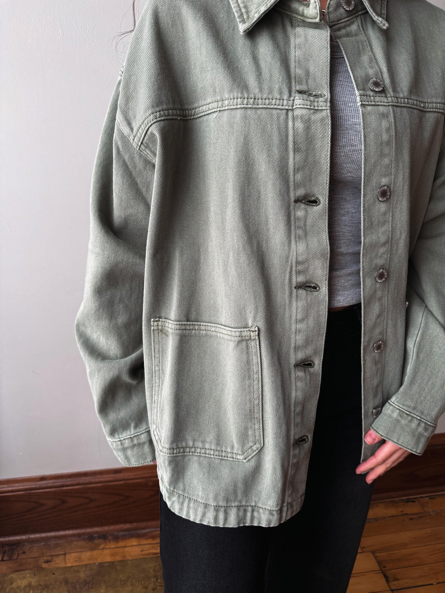 A person wearing an oversized light sage green washed denim jacket over a gray top and black pants, standing indoors against a plain wall and wooden floor.