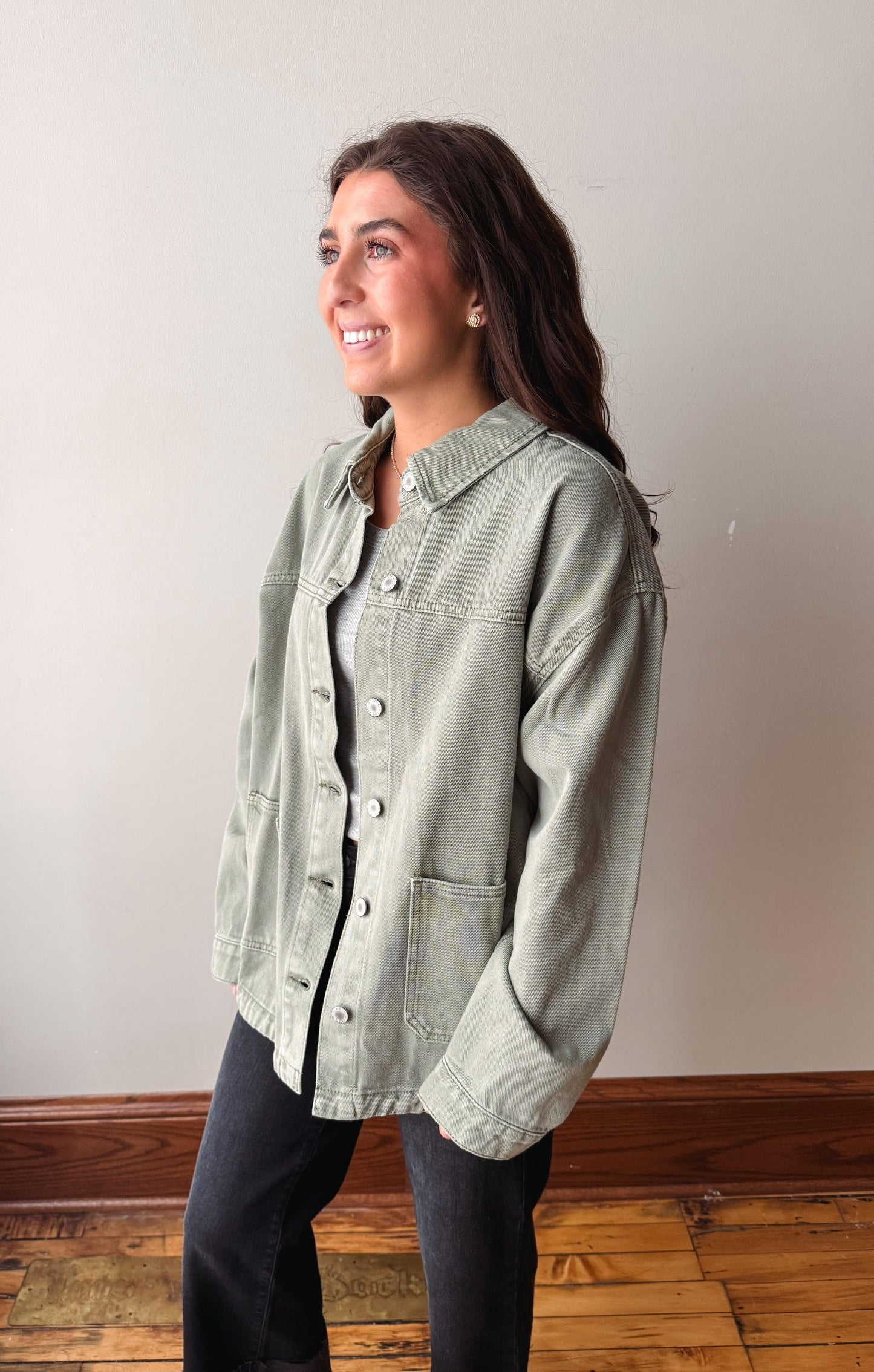 A woman wearing a light sage green denim jacket over a black top and black jeans, standing in a room with a beige wall and wooden floor.