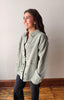 A woman wearing a light sage green denim jacket over a black top and black jeans, standing in a room with a beige wall and wooden floor.