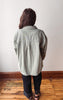 A person viewed from behind wearing an oversized light sage green denim shirt jacket, black pants, and sandals, standing on a wooden floor against a plain off-white wall.