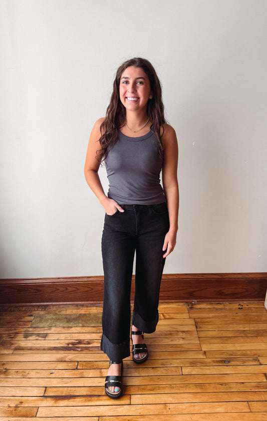 A woman modeling a dark gray ribbed tank top and black wide-leg cropped jeans with black sandals, standing on a wooden floor against a plain off-white wall.