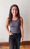 A smiling young woman with long brown hair stands indoors wearing a fitted dark gray ribbed tank top and black jeans against a plain off-white wall with a wooden floor visible.
