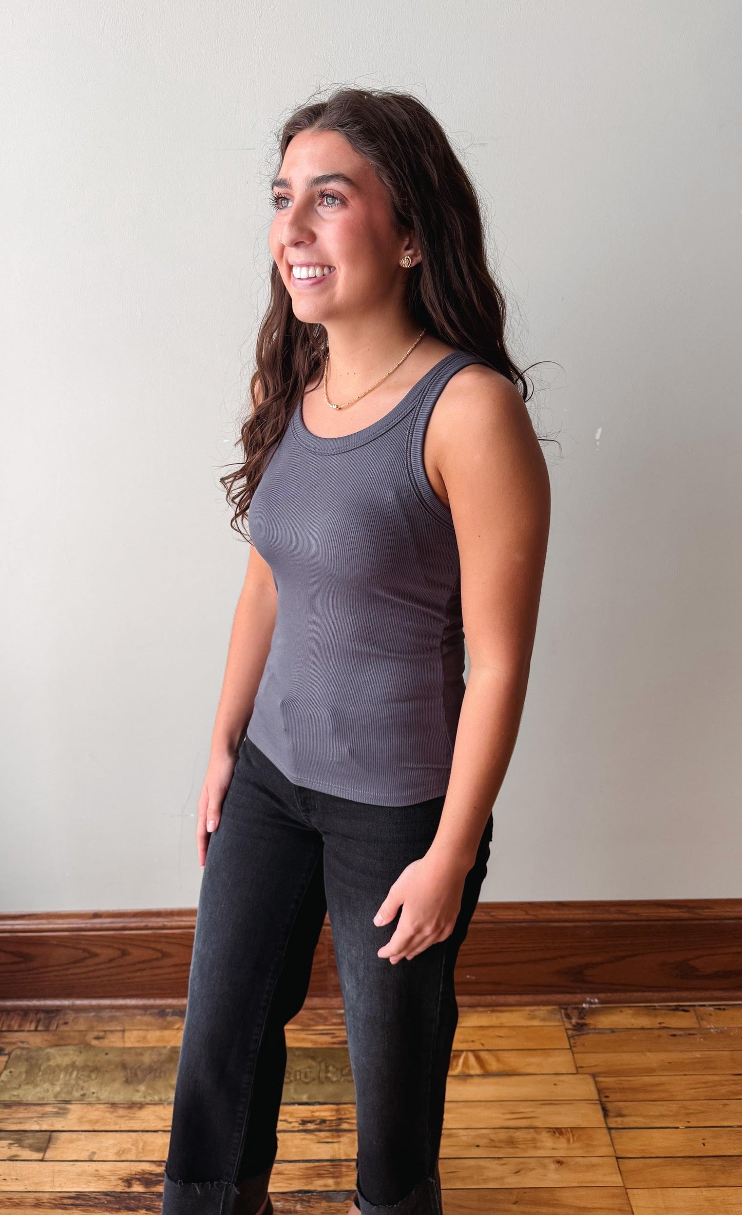 A woman wearing a dark gray ribbed tank top and black jeans, standing in a room with a light gray wall and wooden floor.