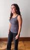 A woman wearing a dark gray ribbed tank top and black jeans, standing in a room with a light gray wall and wooden floor.