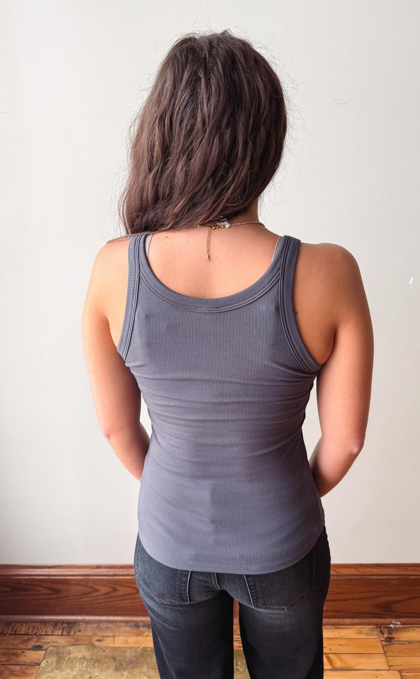 A woman viewed from behind wearing a dark gray ribbed tank top and dark jeans, standing against a plain off-white wall with a wooden baseboard and floor.