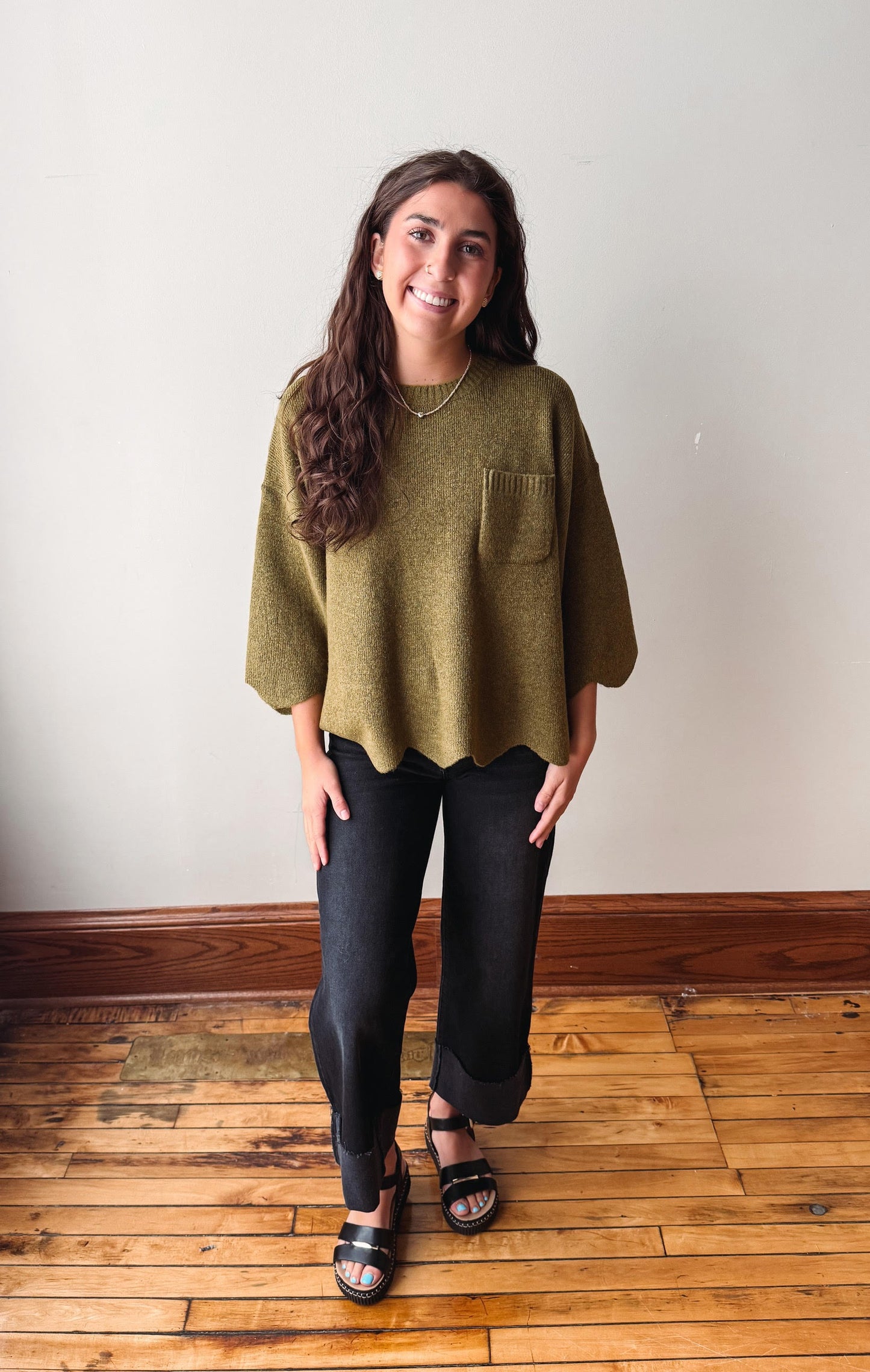 A young woman stands in a room with a light gray wall and wooden floor, wearing a loose olive green knit top with a chest pocket, black wide-leg cropped pants, and black slide sandals.