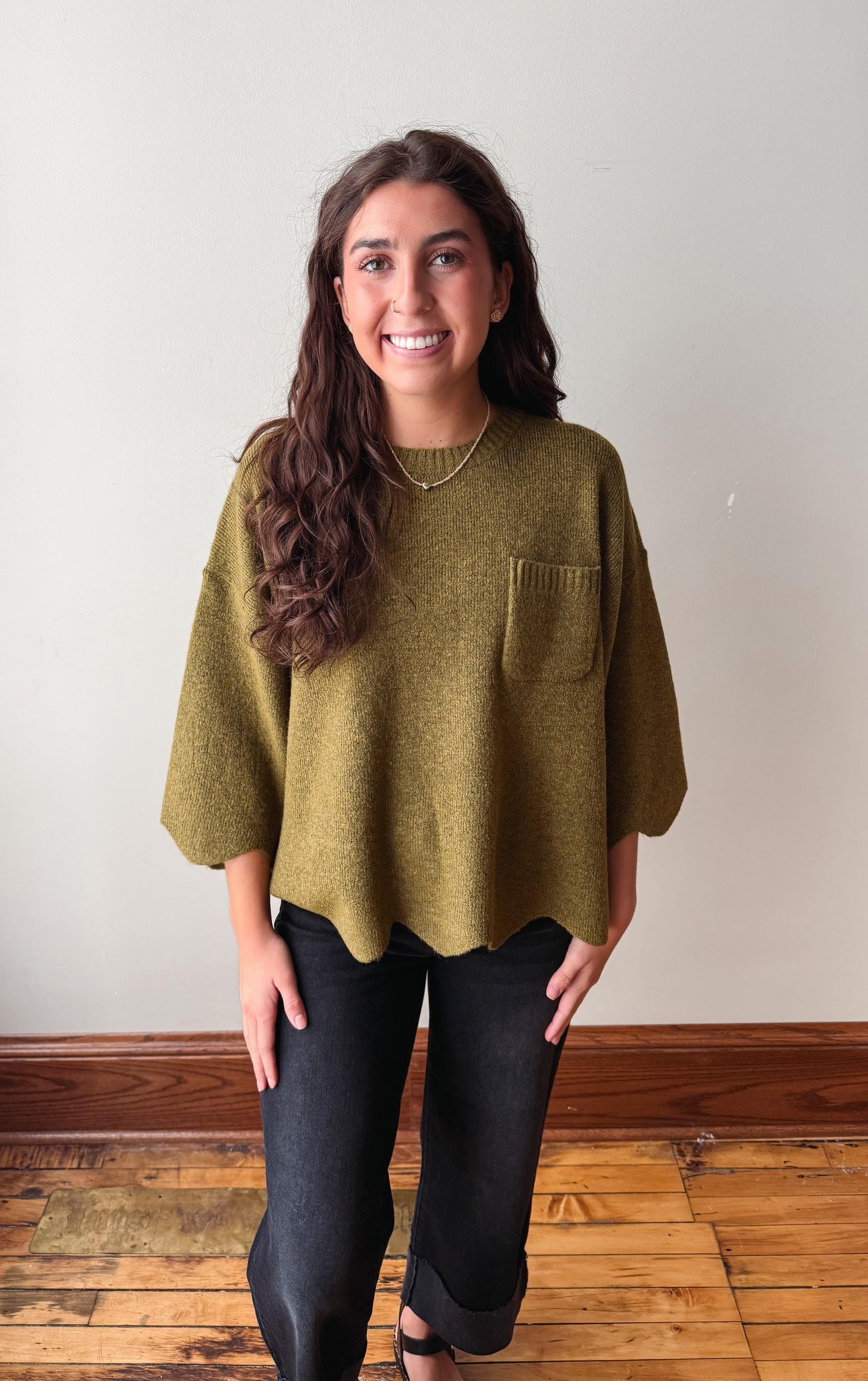 A woman with long brown hair stands indoors wearing an olive green knit top with elbow-length sleeves, a chest pocket, and black pants against a plain off-white wall and wood floor.