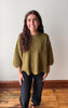 A woman with long brown hair stands indoors wearing an olive green knit top with elbow-length sleeves, a chest pocket, and black pants against a plain off-white wall and wood floor.