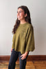 A woman wearing a textured olive green knit sweater with a chest pocket and black pants, standing in a room with a light gray wall and wooden floor.