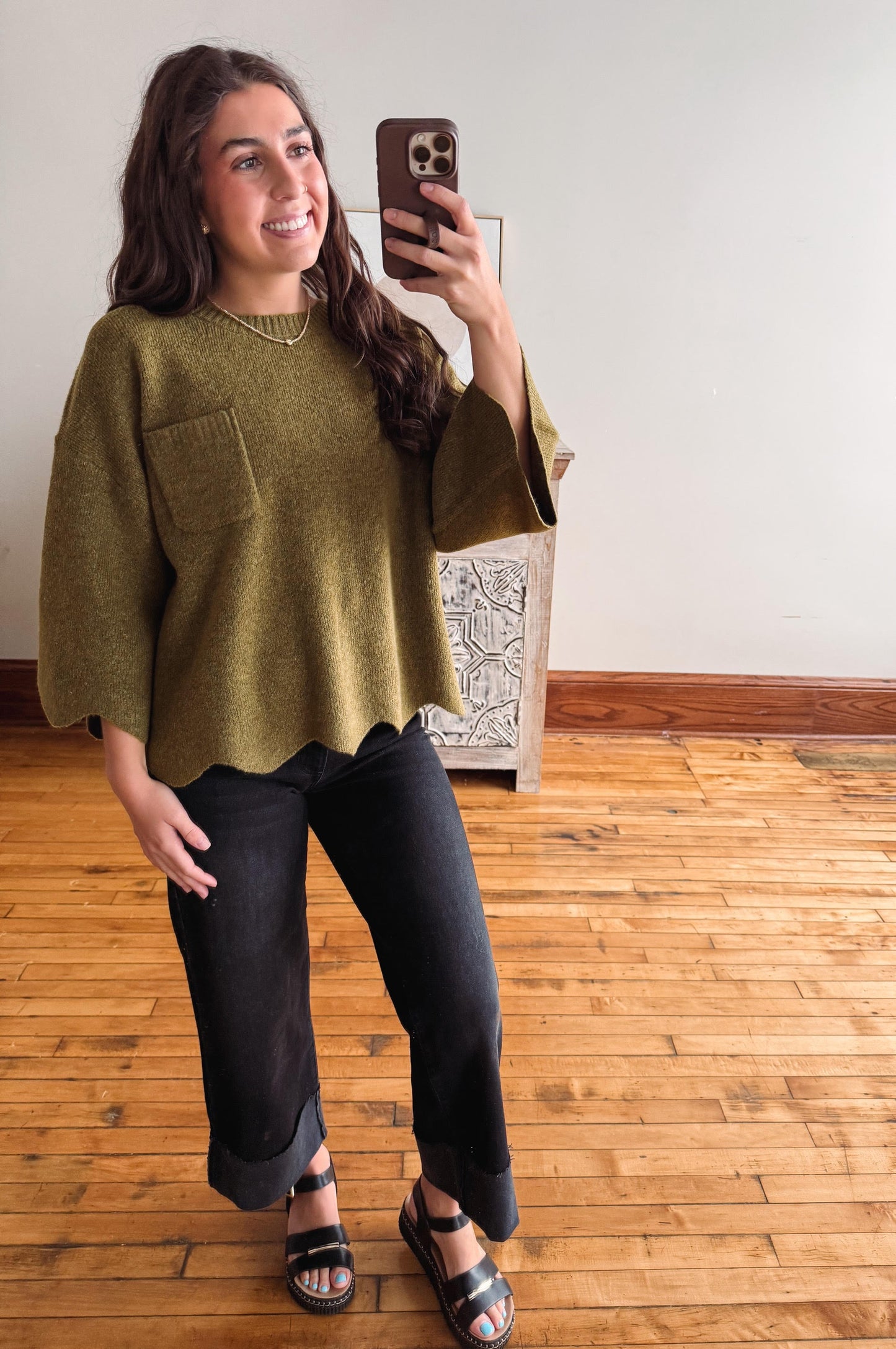 A woman takes a mirror selfie wearing a loose, olive green knit top with wide bell sleeves and a chest pocket, paired with black jeans and black platform sandals, in an indoor room with wooden floors.
