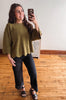 A woman takes a mirror selfie wearing a loose, olive green knit top with wide bell sleeves and a chest pocket, paired with black jeans and black platform sandals, in an indoor room with wooden floors.