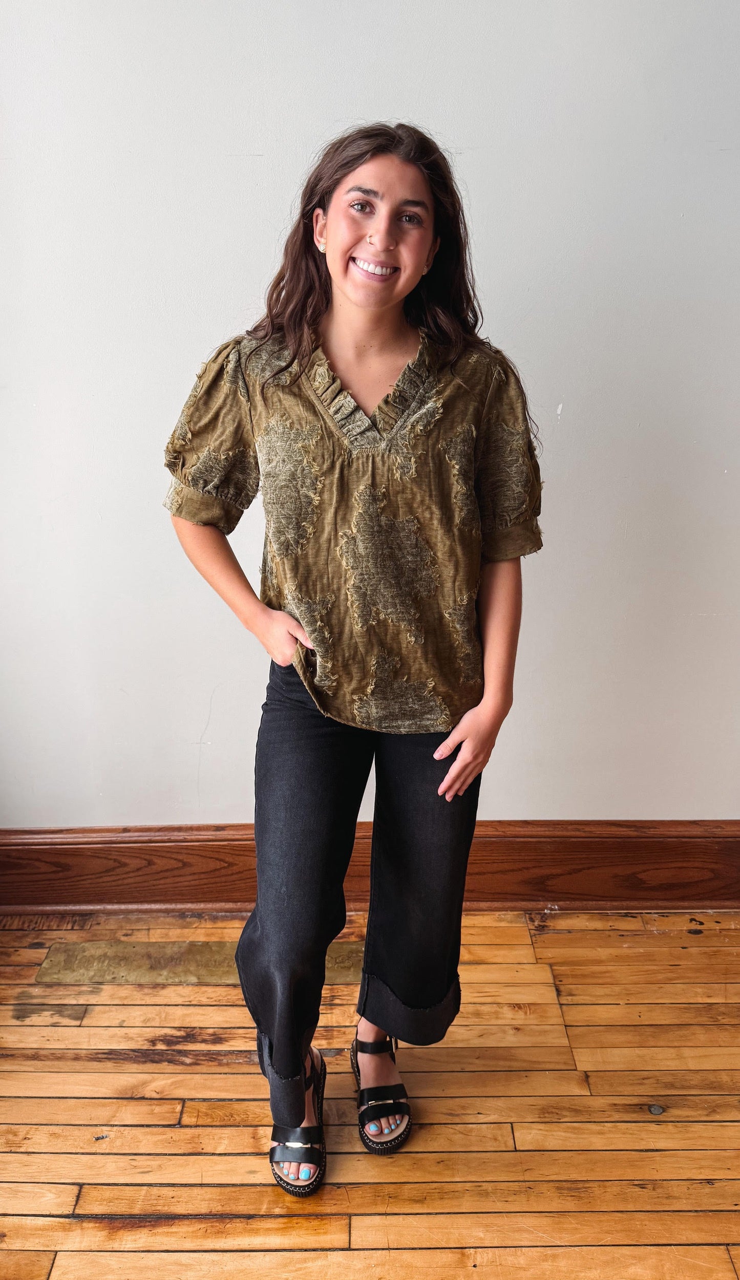 A woman modeling a short-sleeve textured blouse in olive green and gold, paired with black pants and black sandals, standing on a wooden floor against a plain white wall.