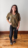A woman modeling a short-sleeve textured blouse in olive green and gold, paired with black pants and black sandals, standing on a wooden floor against a plain white wall.