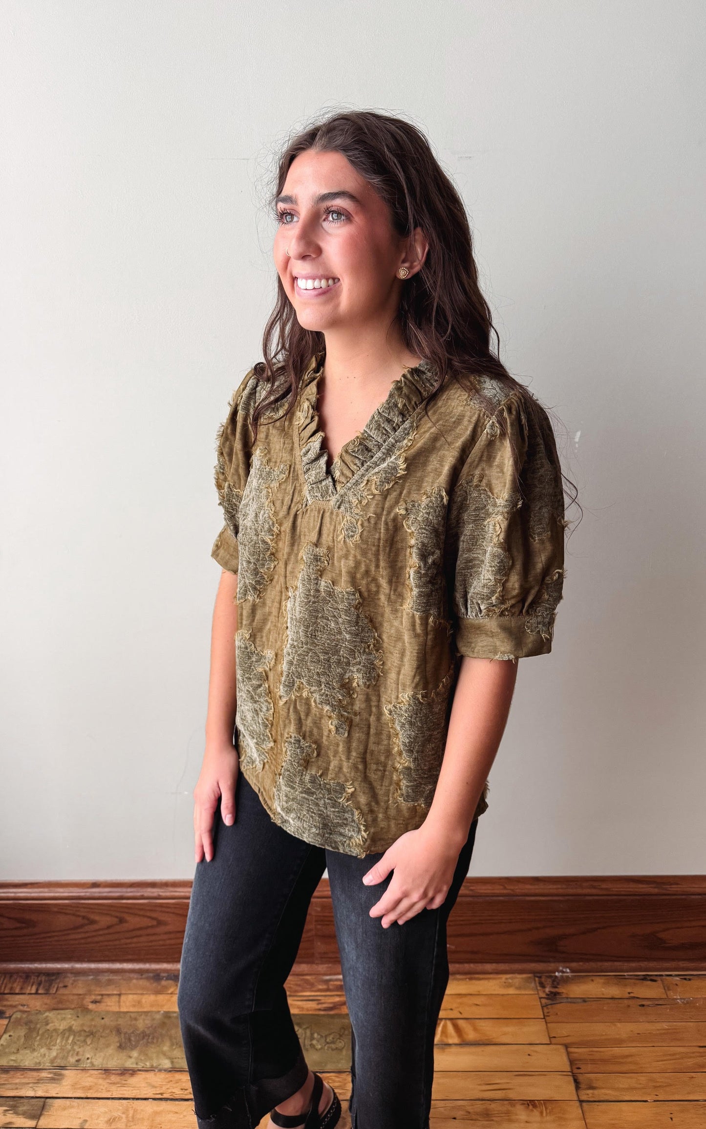 A woman modeling a short-sleeve V-neck blouse with a textured, distressed olive green and brown pattern, paired with black jeans, standing against a plain off-white wall with wooden trim and a wood floor.