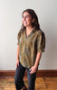 A woman modeling a short-sleeve V-neck blouse with a textured, distressed olive green and brown pattern, paired with black jeans, standing against a plain off-white wall with wooden trim and a wood floor.