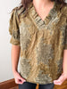 A person wearing a distressed olive green and beige patterned short-sleeve blouse with a ruffled V-neckline, paired with dark jeans.