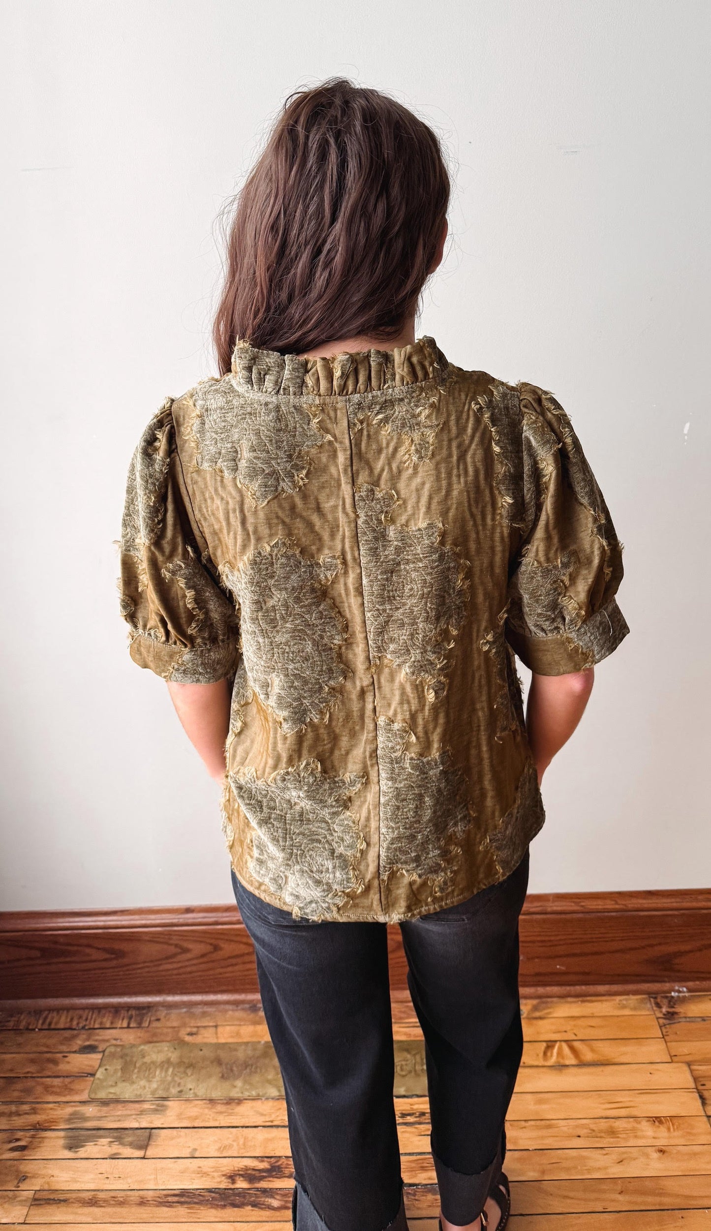 A person viewed from behind wearing a textured, patterned short-sleeve blouse in olive green and gold tones, paired with black pants, standing on a wooden floor against a plain white wall.