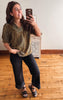 A woman takes a mirror selfie in a room with hardwood floors, wearing a short-sleeved olive green and gold patterned blouse, black wide-leg cropped pants, and black sandals.