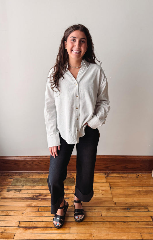 A woman stands in a room with a light gray wall and wooden floor, wearing a light beige long-sleeve button-up shirt, black wide-leg pants, and black sandals.