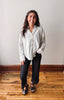 A woman stands in a room with a light gray wall and wooden floor, wearing a light beige long-sleeve button-up shirt, black wide-leg pants, and black sandals.