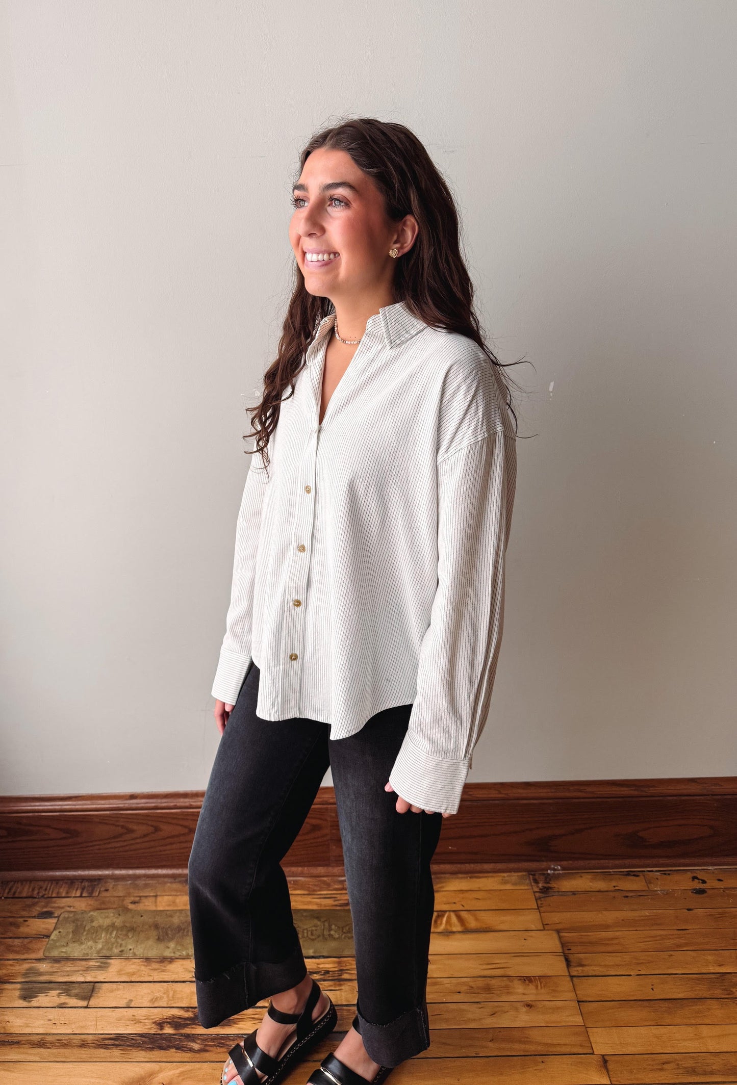 A woman wearing a white long-sleeve button-up shirt and black pants, standing in a room with a light gray wall and wooden floor.