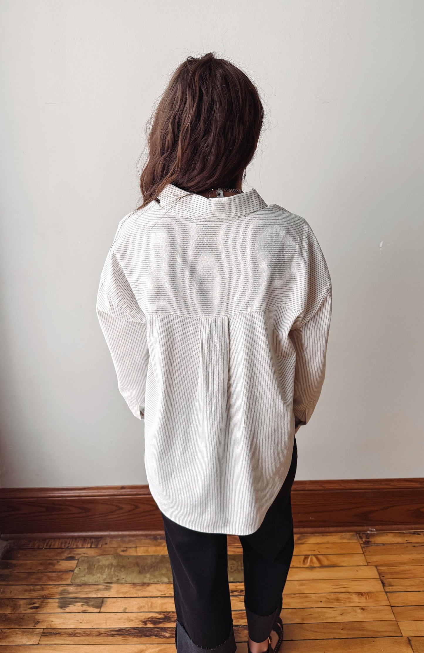 A person viewed from behind wearing a loose, off-white long-sleeve button-up shirt and black pants, standing on a wooden floor against a plain light wall.