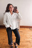 A woman takes a mirror selfie wearing a white long-sleeve button-down shirt with gold buttons and black wide-leg pants, standing on a wooden floor against a plain wall.