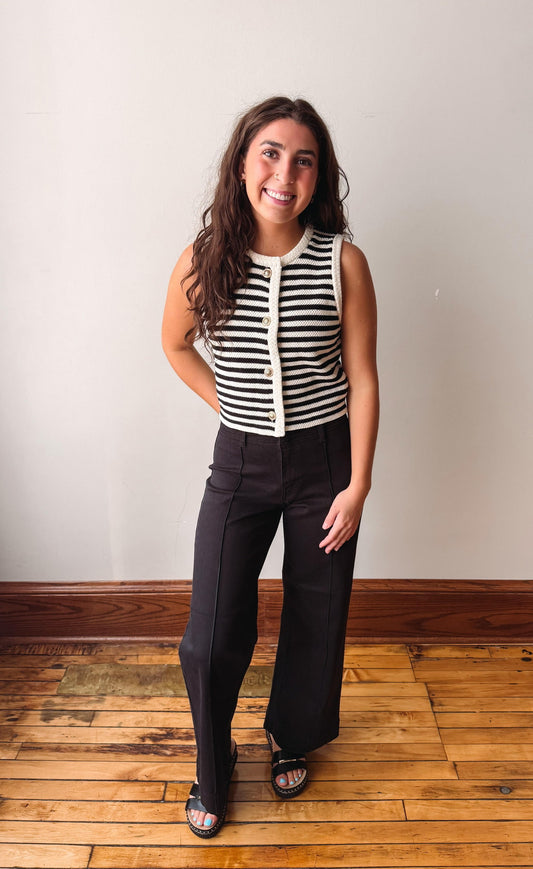 A woman modeling a sleeveless black and white striped top with a cream button placket, paired with black wide-leg pants and black slide sandals, standing on a wooden floor against a plain off-white wall.