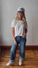A woman stands in a room with a light gray wall and wooden floor, wearing a white short-sleeve t-shirt with a small gold chest logo, blue jeans with rolled cuffs, white sneakers, and a white baseball cap with blue lettering.