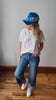 A woman wearing a blue baseball cap with 'USA STATE' printed on it, a white t-shirt, blue jeans, and white sneakers, standing on a wooden floor against a plain wall.