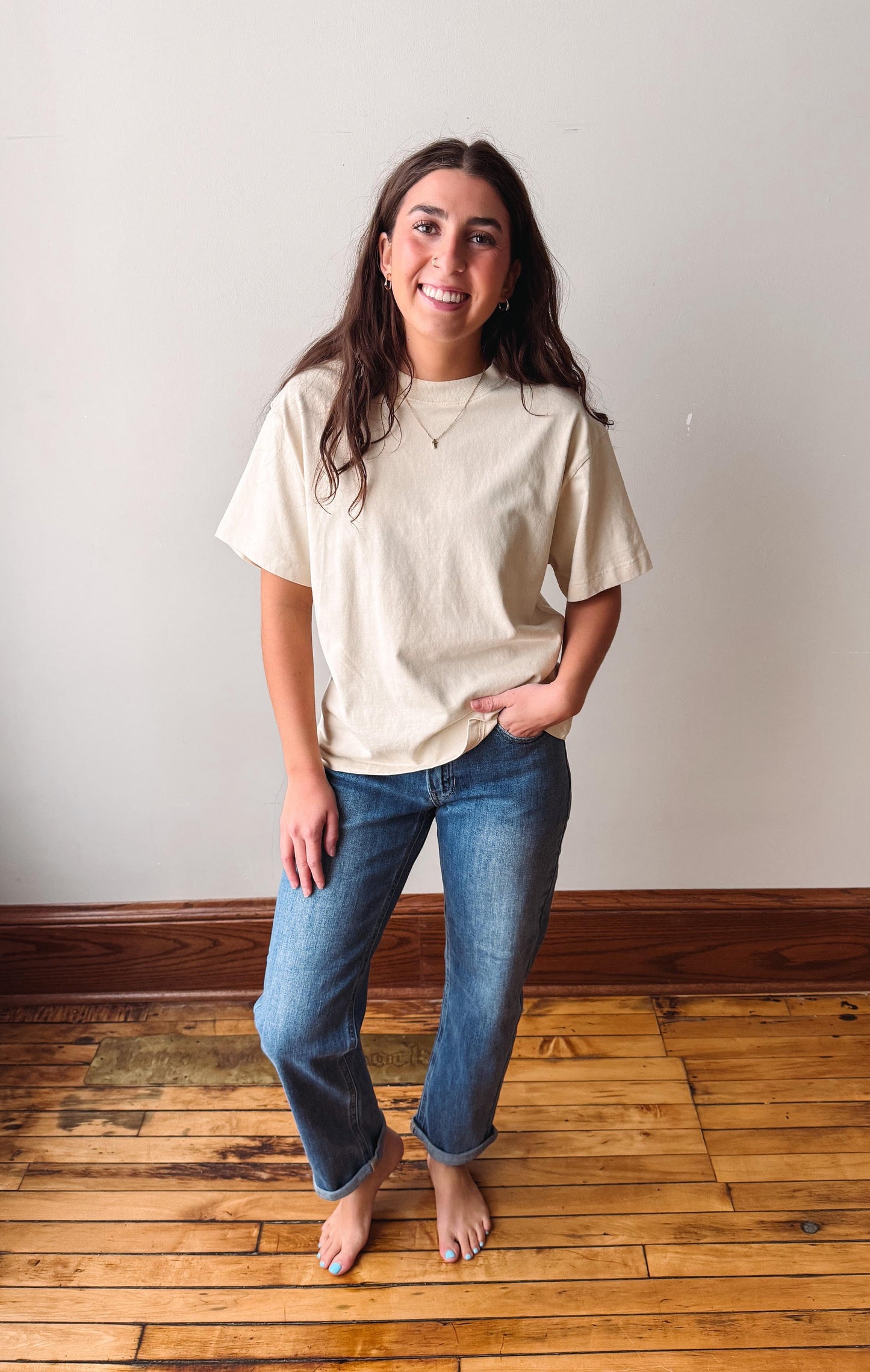 A woman stands barefoot on a wooden floor against a plain off-white wall, wearing a loose beige short-sleeve t-shirt and blue jeans.