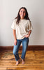 A woman stands barefoot on a wooden floor against a plain off-white wall, wearing a loose beige short-sleeve t-shirt and blue jeans.