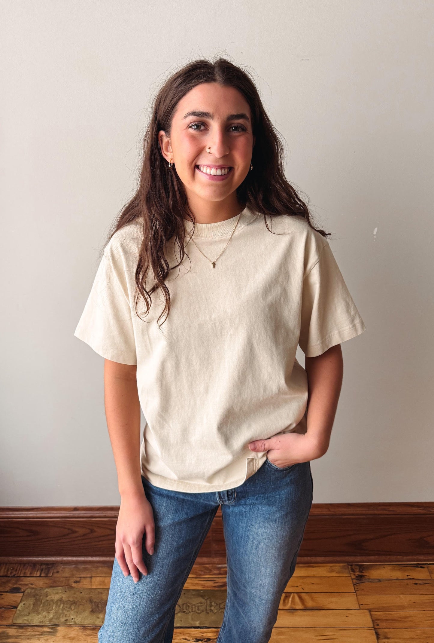 A smiling young woman with long dark hair stands indoors wearing an oversized cream-colored t-shirt and blue jeans against a plain light wall with a wooden floor.