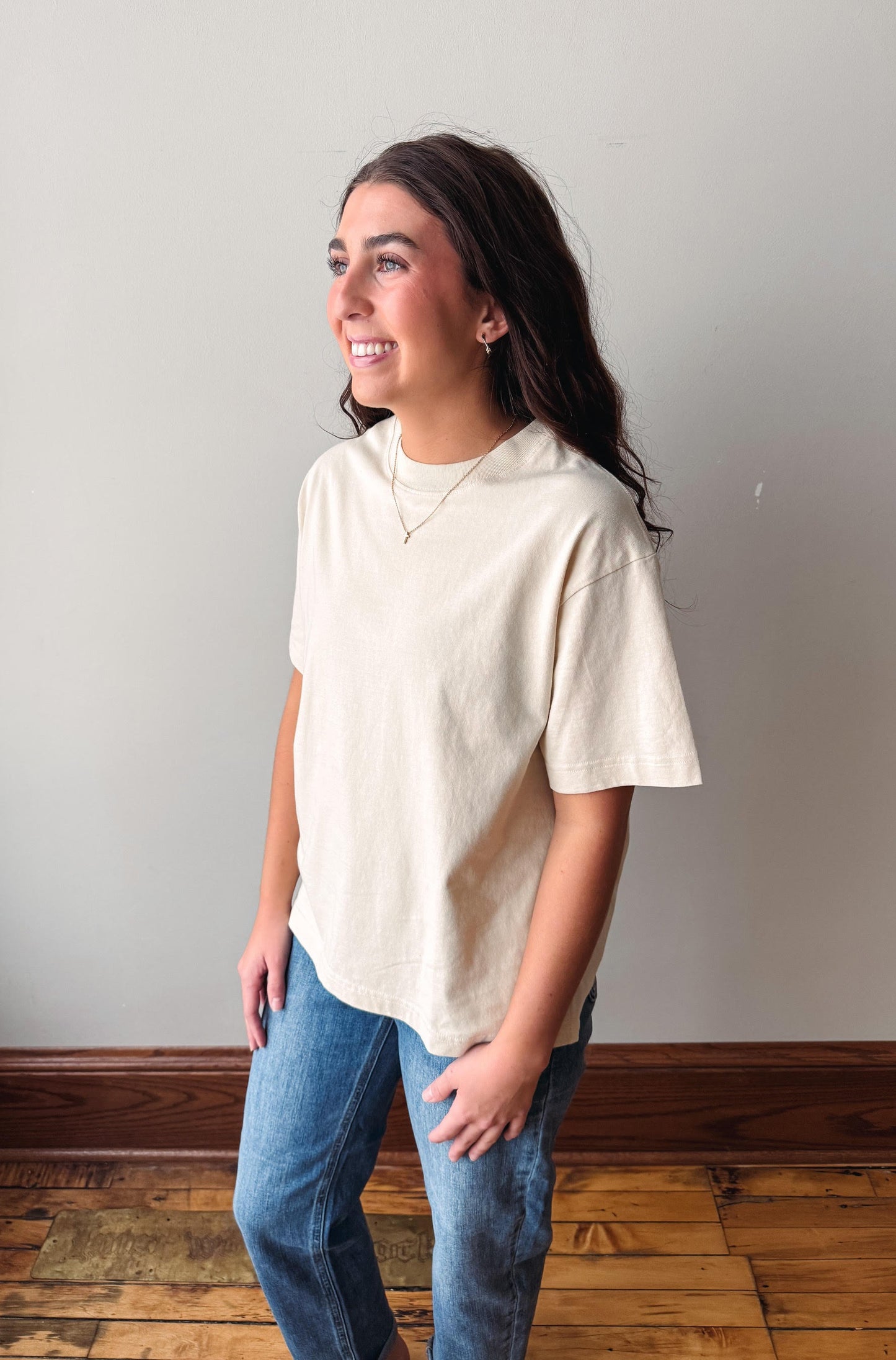 A woman wearing a cream-colored short-sleeve t-shirt and blue jeans, standing in a room with a light gray wall and wooden floor.
