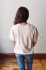 A person viewed from behind wearing an oversized off-white t-shirt and blue jeans, standing on a wooden floor against a plain light gray wall.