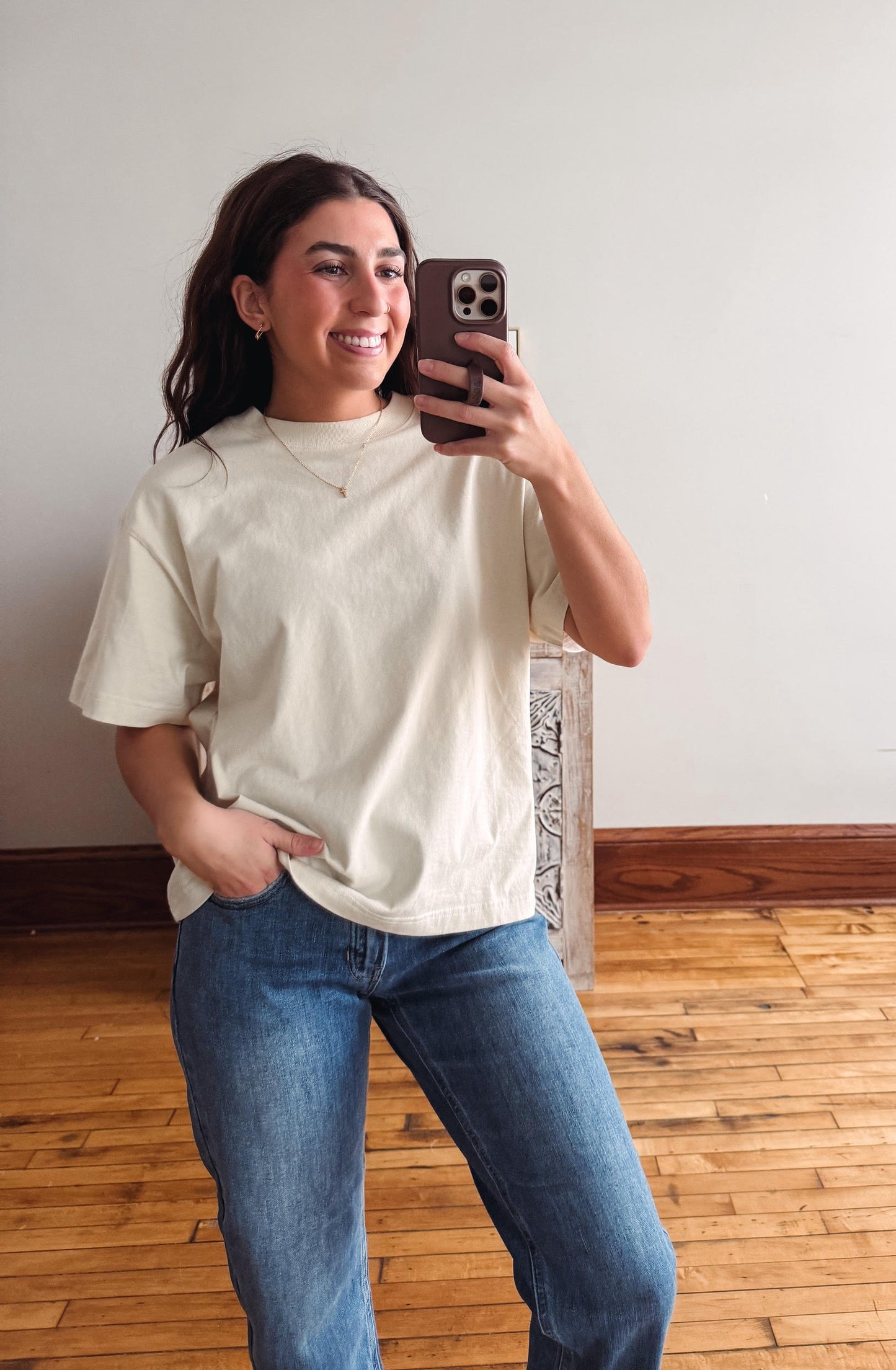 A woman taking a mirror selfie wearing a cream-colored short-sleeve t-shirt and blue jeans in a room with a light gray wall and wooden floor.