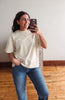 A woman taking a mirror selfie wearing a cream-colored short-sleeve t-shirt and blue jeans in a room with a light gray wall and wooden floor.