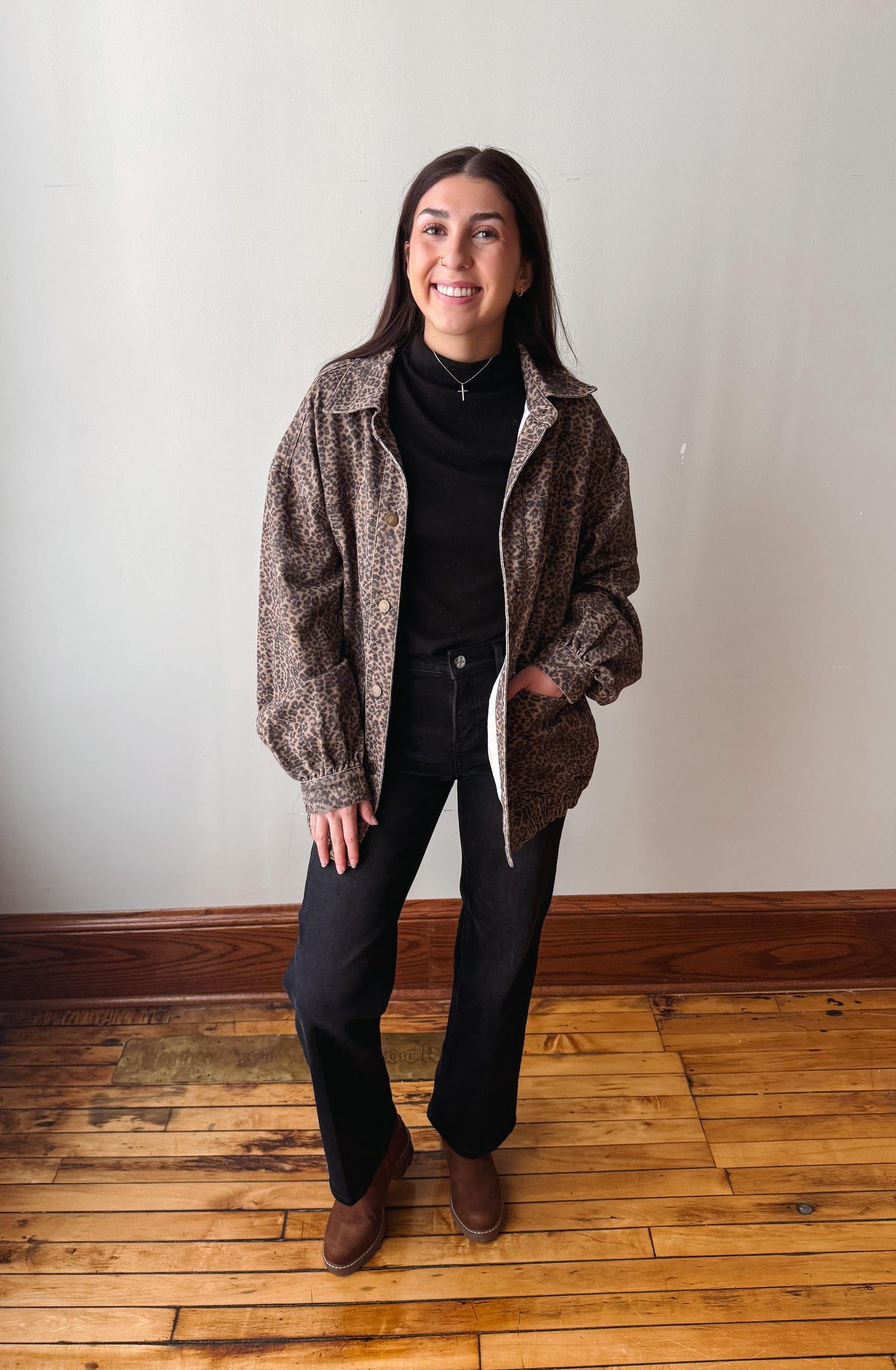 A woman stands in a room with a light gray wall and wooden floor, wearing a brown and black patterned zip-up jacket over a black turtleneck, black pants, and brown boots.