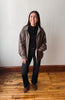 A woman stands in a room with a light gray wall and wooden floor, wearing a brown and black patterned zip-up jacket over a black turtleneck, black pants, and brown boots.