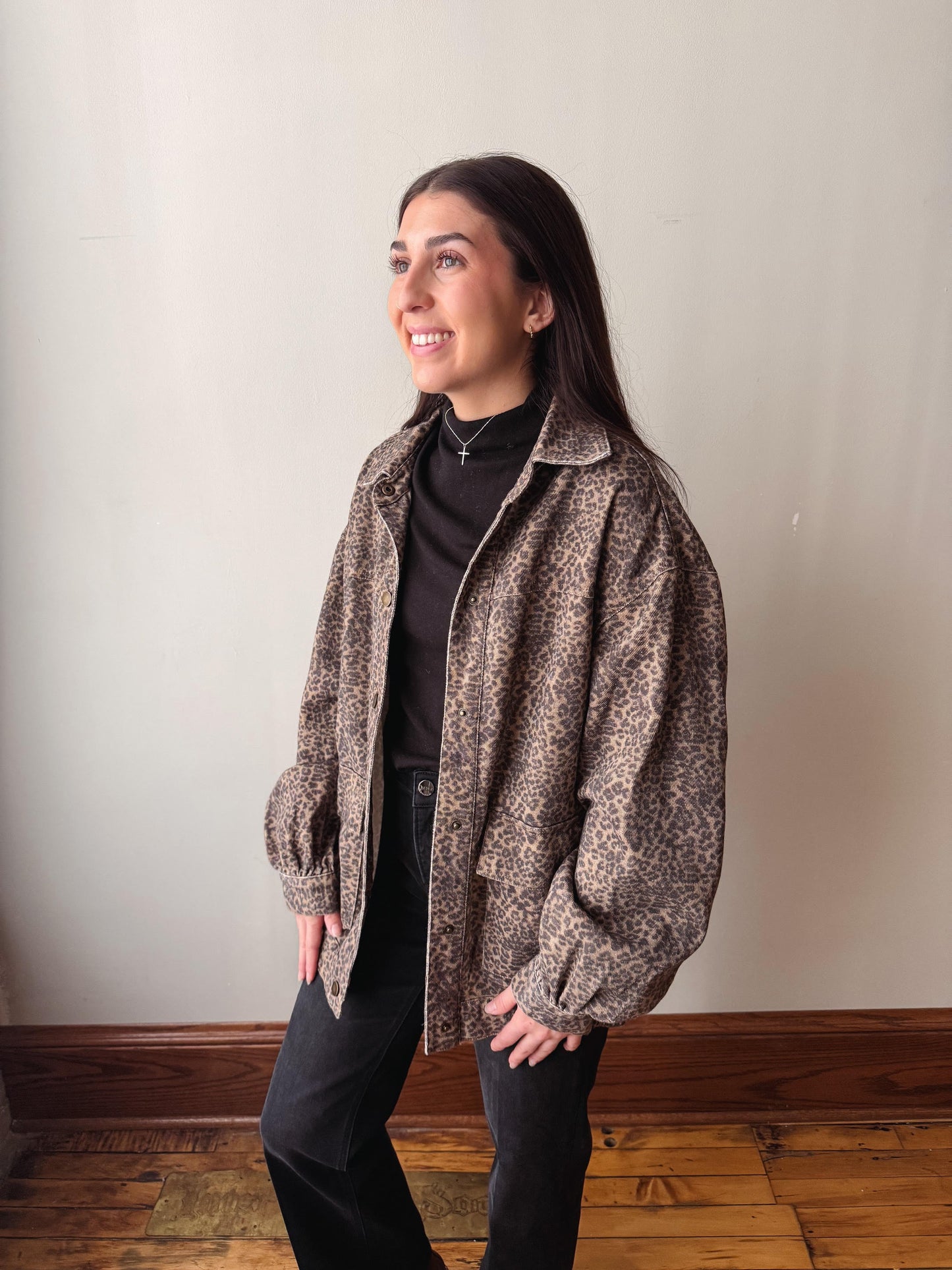 A woman wearing a brown and beige patterned zip-up jacket over a black turtleneck and black pants, standing in an indoor setting with a light-colored wall and wooden floor.