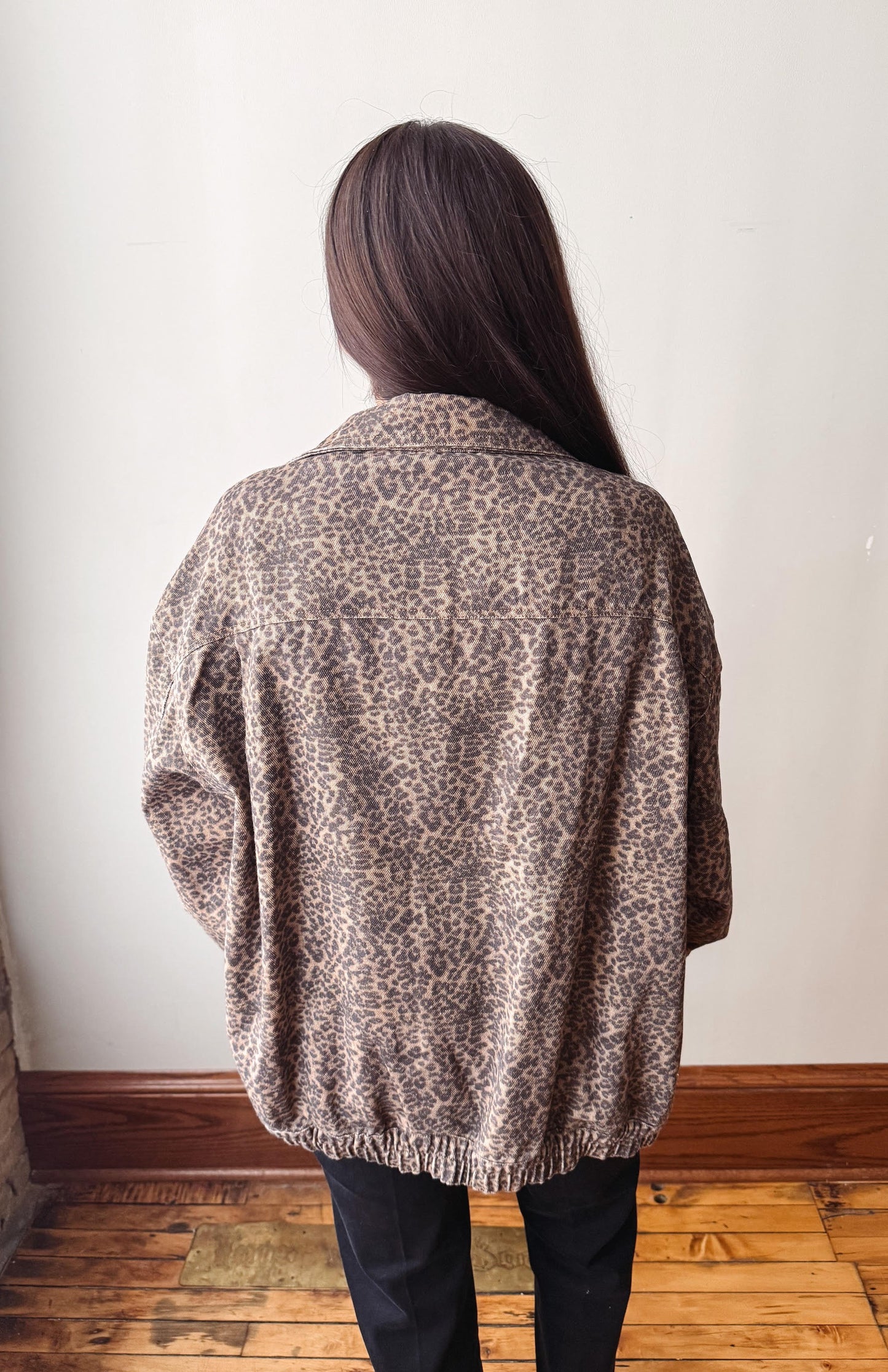A person viewed from behind wearing a brown and beige leopard-print cardigan or jacket, paired with black pants, standing in front of a plain off-white wall on a wooden floor.