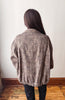A person viewed from behind wearing a brown and beige leopard-print cardigan or jacket, paired with black pants, standing in front of a plain off-white wall on a wooden floor.