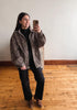 A woman takes a mirror selfie wearing a brown and black patterned sherpa jacket over a black top and black pants, standing on a wooden floor against a plain wall.