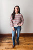 A young woman stands in a room with a light gray wall and wooden floor, wearing a red, black, and white striped knit sweater, blue jeans, and brown boots.
