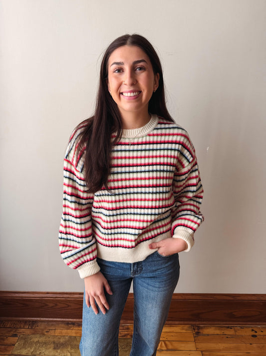 A woman with long dark hair stands indoors wearing a knit sweater with horizontal red, black, and off-white stripes, paired with blue jeans, against a plain beige wall.