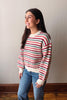 A woman wearing a striped knit sweater with red, black, and off-white horizontal stripes, paired with blue jeans, standing against a plain beige wall.